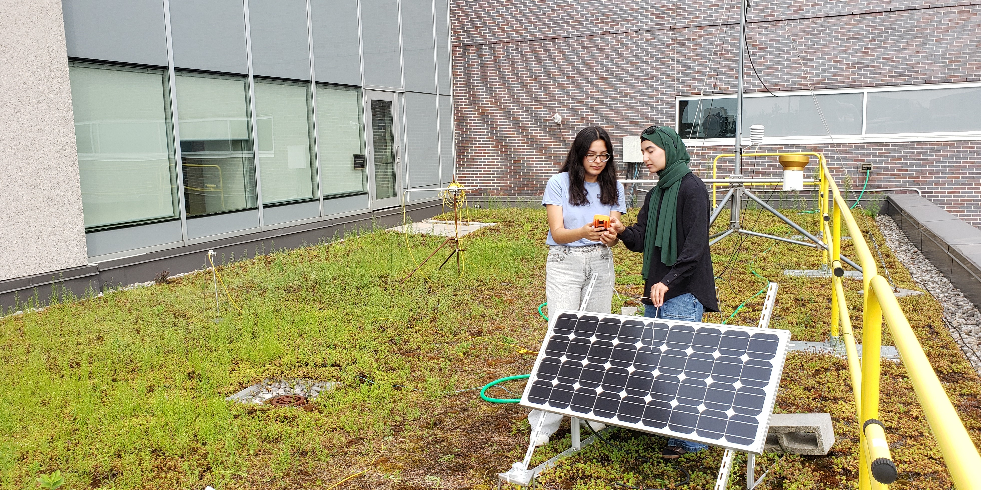 Sevda and Juliana using a multimeter to complete measurements on a small solar panel surrounded by a sedum-covered green roof.